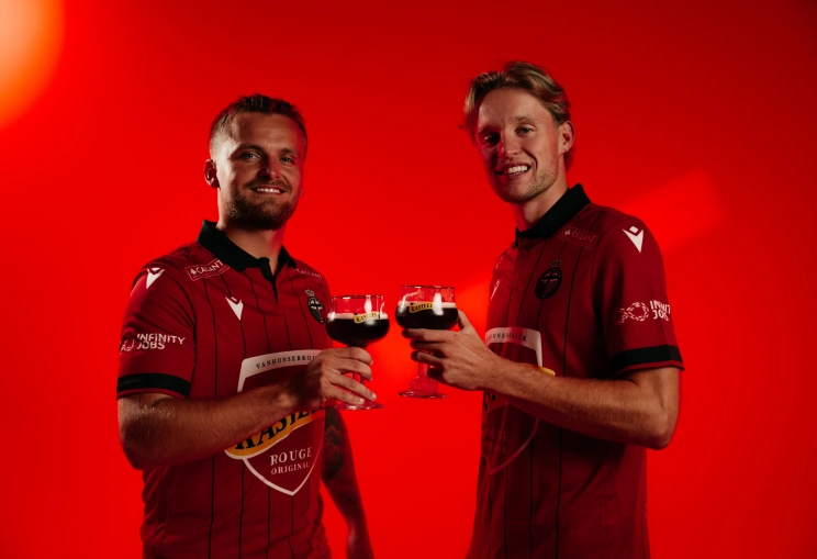 Mandel United uitrusting 2026-2027 met spelers Liam Prez en Jules Desot met elk een glas Kasteel Rouge